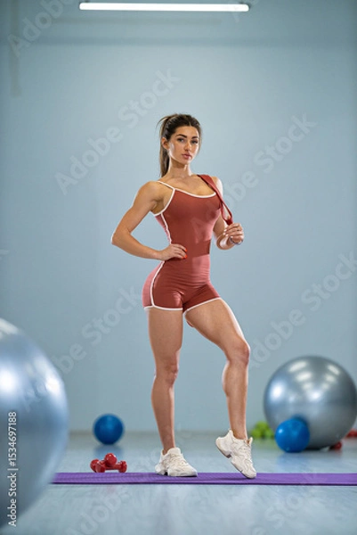 Fototapeta girl stands confidently in a gym, showcasing her sporty attire She strikes a pose while surrounded by gym equipment, including exercise balls and weights, emphasizing her athleticism
