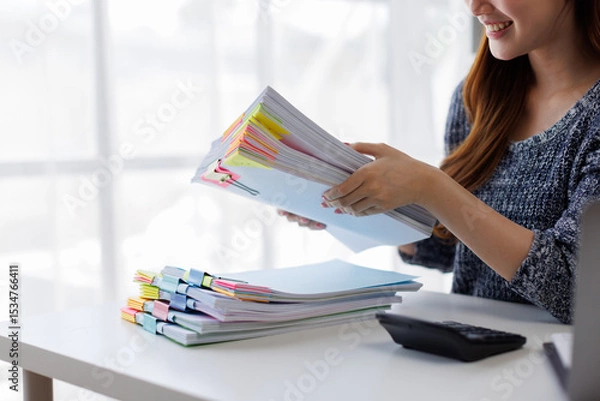 Obraz Image of young Asian business woman working working in Stacks of paper files for searching and checking unfinished document achieves on folders papers at office
