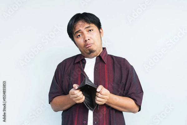 Obraz Asian Indonesian man showing his empty wallet with sad and worried expression. Isolated in gray background.
