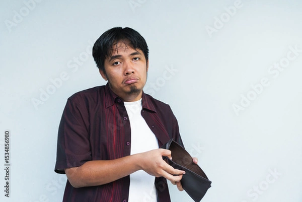 Obraz Asian Indonesian man showing his empty wallet with sad and worried expression. Isolated in gray background.