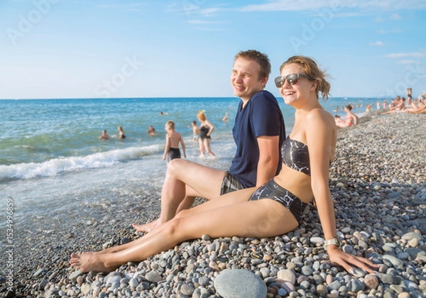 Fototapeta A young smiling guy and a girl are sitting by the sea on a crowded pebble beach against the background of the blue sea and clear sky on a bright sunny day. Side view