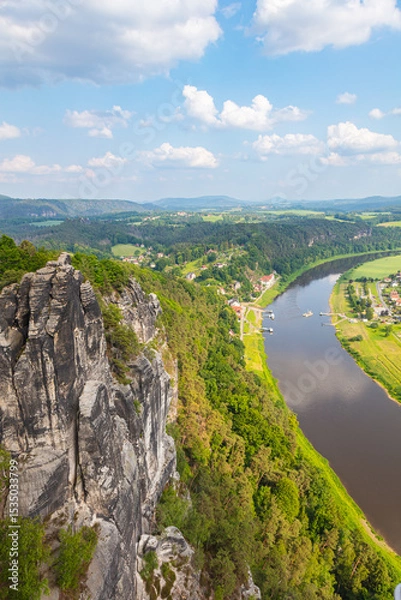 Obraz Scenic panoramic view from the Bastei cliffs over the Elbe River valley with sandstone rocks, lush forests, rural village rooftops, distant mountain ranges under a vibrant summer sky. Saxony, Germany