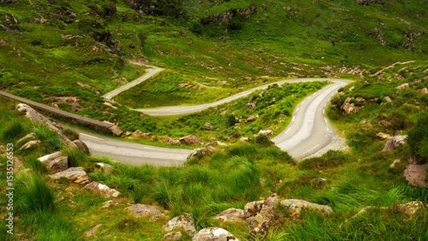 Fototapeta A narrow road winds sharply through a verdant, hilly landscape. Rocks and tall grass grow alongside the road. The Gap of Dunloe shows its beautiful scenery.
