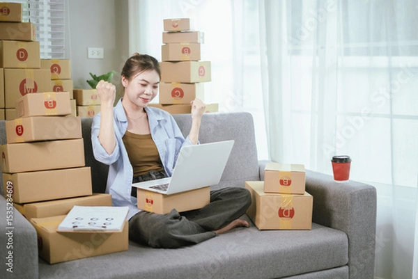 Fototapeta An Asian female entrepreneur is preparing parcels and checking online orders of her products to ship to customers using a laptop at home. A concept of online shopping and a small startup SME business.
