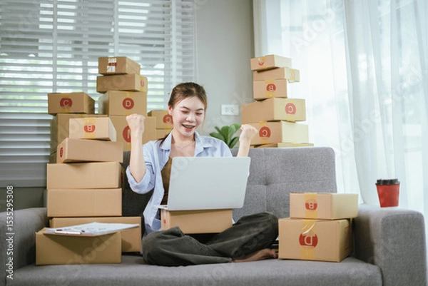 Fototapeta An Asian female entrepreneur is preparing parcels and checking online orders of her products to ship to customers using a laptop at home. A concept of online shopping and a small startup SME business.