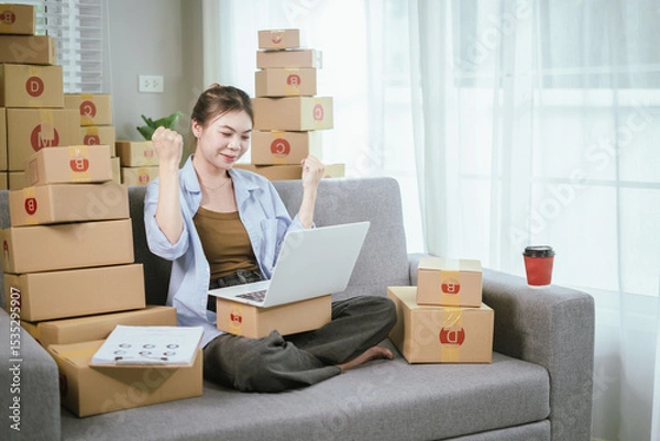 Fototapeta An Asian female entrepreneur is preparing parcels and checking online orders of her products to ship to customers using a laptop at home. A concept of online shopping and a small startup SME business.