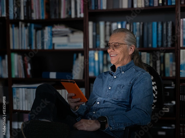 Fototapeta Smiling man enjoying reading on a tablet in a private library