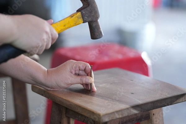 Obraz a person using a hammer to drive a nail into a wooden surface, with one hand holding the nail and the other gripping the hammer. This scene represents woodworking or DIY repair work