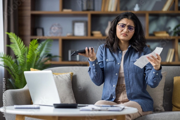 Fototapeta Portrait of upset young Indian woman sitting on sofa at home, holding documents, bills and mobile phone, spreading her arms at camera in disappointment