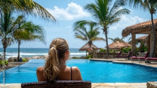 Fototapeta Reclining comfortably by a sparkling pool at a luxury beach resort, a young woman enjoys the serenity of the moment while taking in the stunning oceanfront views.