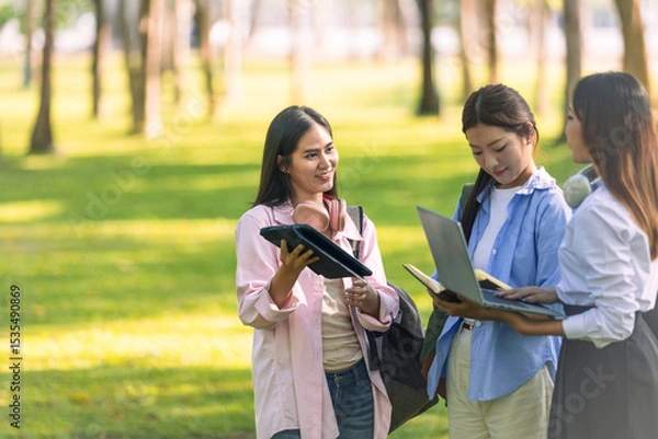 Obraz Group of Asian college students standing on green grass and discussing with laptop and books. Concept of teamwork, education, and outdoor learning in a natural environment.