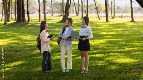Obraz Group of Asian college students standing on green grass and discussing with laptop and books. Concept of teamwork, education, and outdoor learning in a natural environment.