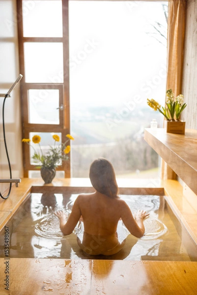 Fototapeta A person relaxes in a wooden hot tub facing a panoramic countryside view. The warm steam rises, surrounded by flowers and soft morning light through the large window