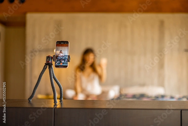 Fototapeta A smartphone on a tripod captures a woman in pajamas creating video content. Great for illustrating influencer work, home blogging, or remote lifestyle content creation