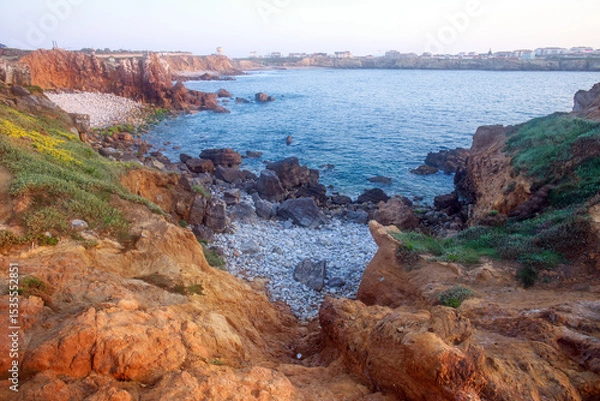 Fototapeta Sunny rocky coastline of Atlantic Ocean in the rays of setting sun, Papoa Island, Peniche, Portugal