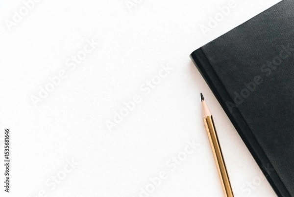 Fototapeta A sharpened pencil lies next to a closed black notebook on a clean white surface.