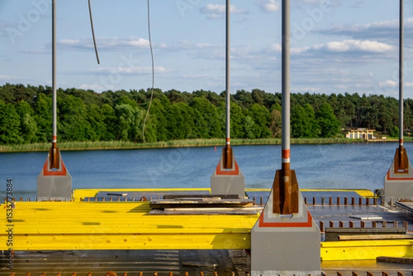 Fototapeta Section of a bridge under construction over water, with visible yellow formwork and metal support elements. A scenic background with a lake and green forest.