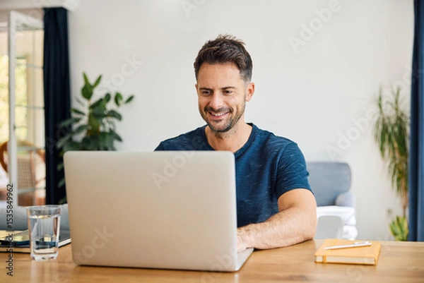 Fototapeta Smiling man sitting at desk with laptop at home