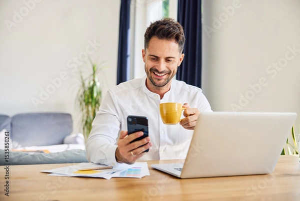 Fototapeta Smiling freelancer using mobile phone at home while working on laptop