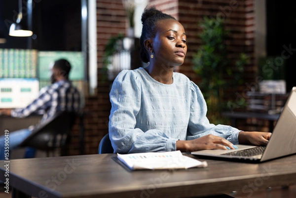 Obraz Young businesswoman with clipboard nearby, working late at night, analyzing financial data on her laptop. A blurred male coworker in the background focuses on multiple monitors.