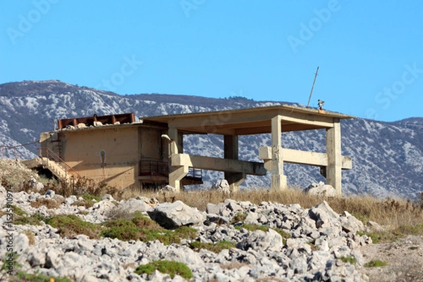 Fototapeta A partially constructed or abandoned concrete building frame with exposed beams and columns sits on a rocky slope, backed by distant mountains and under a clear blue sky, evoking desolation and unfini