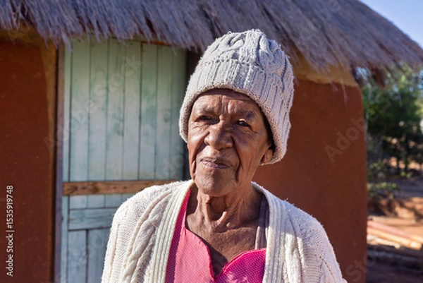 Fototapeta village african old woman standing in the yard in front of a hut with thatched roof, wearing a beanie