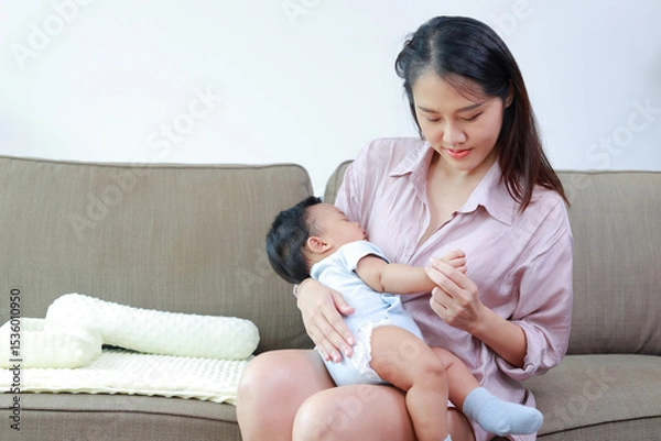 Obraz Asian mother holding her little baby in her arms smiling happily sitting on the sofa at home. Mother and baby bonding helps baby's development. Family concept. Mother's day.