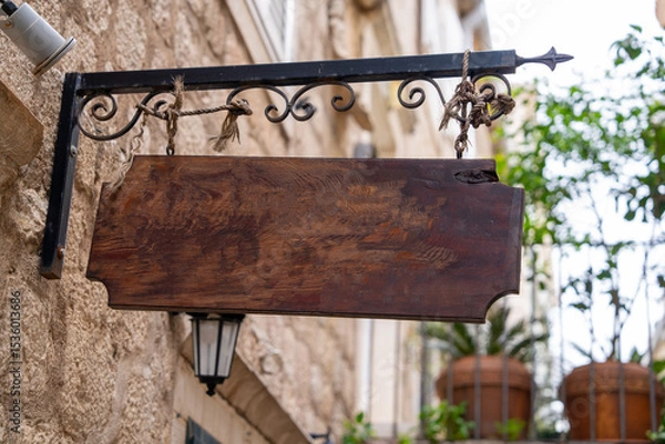 Fototapeta A rustic wooden sign hangs empty from an ornate iron bracket in a charming vintage street. the backdrop features stone walls and potted plants, creating a quaint atmosphere.