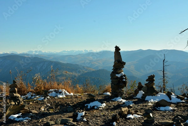 Obraz High pyramids of stones and dry grass sprinkled with first snow on the top of a cliff with a view of mountain ranges on a sunny autumn day.