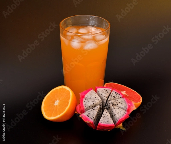 Obraz Fruit juice in a tall glass with ice on a black background, halves of ripe mango, orange and grapefruit nearby.