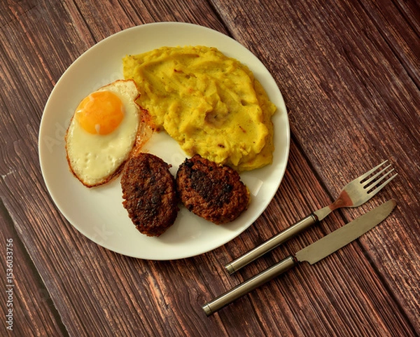 Obraz A large plate with a fried egg, mashed potatoes and two meat patties on a wooden table, cutlery nearby.