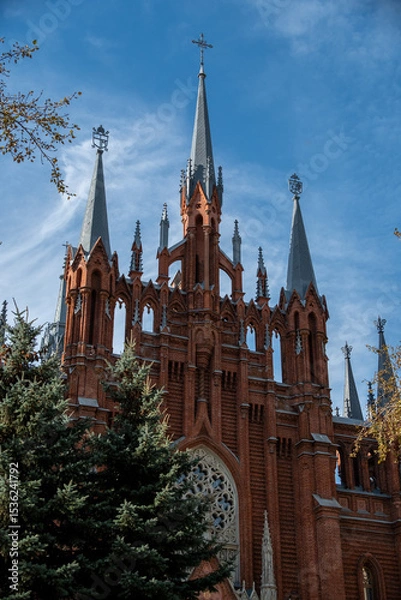Obraz Stunning view of a historic red brick cathedral with elegant spires rising against a clear blue sky. Seen from a tree-lined street with lush greenery enhancing the serene atmosphere.