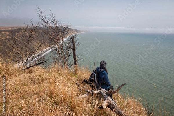 Fototapeta A woman sits on a dead tree trunk on a high seashore and looks into the distance at the sea. Travel and tourism in the Magadan region. The coast of the Sea of ​​Okhotsk. Nature of the Russian Far East