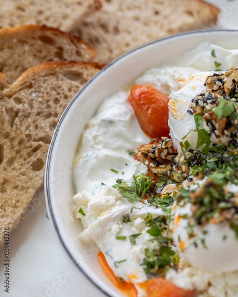 Fototapeta Close-up of a savory breakfast or brunch dish with poached eggs served over creamy yogurt with herbs, cherry tomatoes, sesame seeds, slices of rustic sourdough bread. Mediterranean-inspired meal.