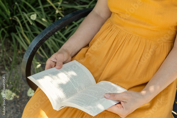 Obraz Close-up of a woman in a mustard yellow dress sitting on a bench and reading a book outdoors with summer garden on the background. Peaceful and mindful moment of relaxation and leisure.
