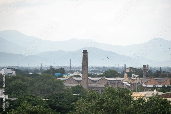 Fototapeta An expansive view of Madurai showcases industrial chimneys among lush greenery, with distant mountains