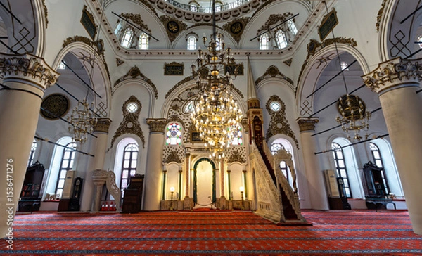 Fototapeta An elaborate chandelier hangs in the center of Hisar Mosque. The historic building has white walls, ornate gold trim, arches, columns, and a red patterned carpet. Located in Izmir, Turkey