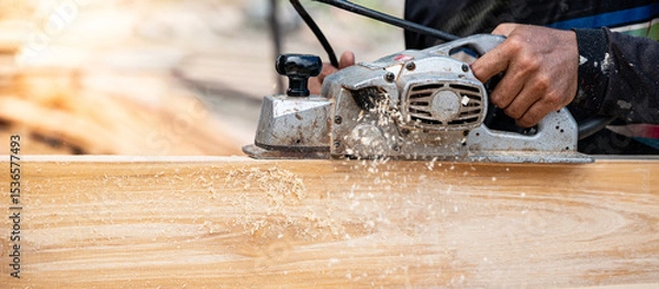 Fototapeta Male carpenter hand using an electric planer on wooden plank at construction site. Woodworker planing timber in his workshop with sawdust spreading around. Lumber industry and carpentry work tool