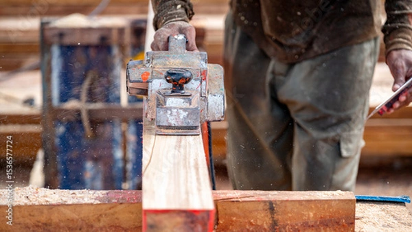Fototapeta Male carpenter hand using an electric planer on wooden plank in workshop. Woodworker planing timber at construction site with sawdust spreading around. Work tool for carpentry. Lumber industry concept
