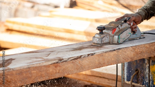 Fototapeta Male carpenter hand using an electric planer on wooden plank in workshop. Woodworker planing timber at construction site with sawdust spreading around. Work tool for carpentry. Lumber industry concept