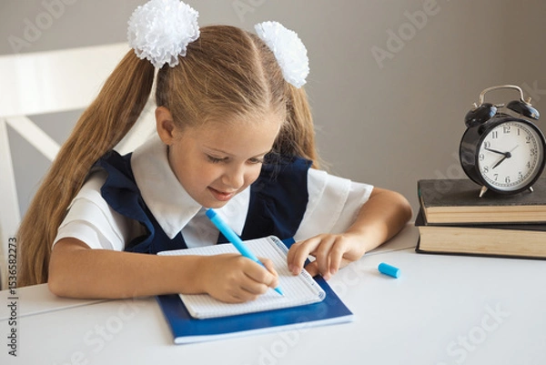 Fototapeta Cute child writing in a notebook with a pen, doing tests. A schoolgirl in uniform sits at a desk and does homework.
