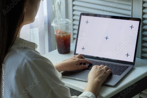 Fototapeta laptop blank white screen mockup. woman using hand typing on keyboard laptop at home for work or entertainment on day time with ice coffee on the table near window with light filtering curtains