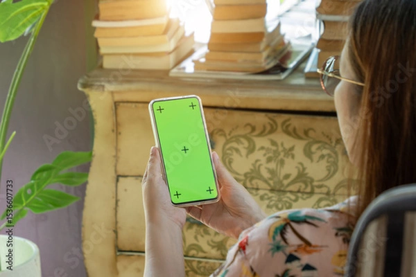 Fototapeta cell phone blank green screen mockup. woman holding mobile phone in hand, using smartphone at living room for work contact or entertainment. room decorate with vintage cabinet, book, Monstera tree
