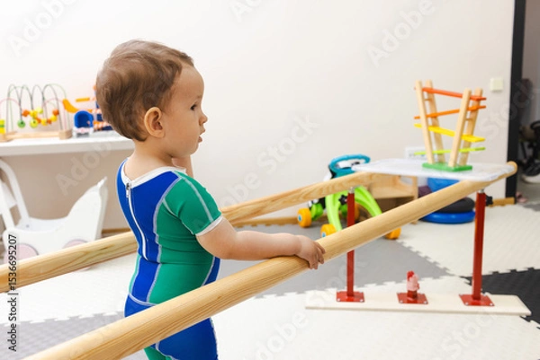 Fototapeta Little male child with movement disorders while participating in physical rehabilitation standing near rehab handrails in vibrant recovery space