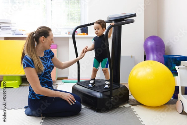Fototapeta Little boy during vibration therapy standing on vibration platform with her assistant in a rehabilitation center
