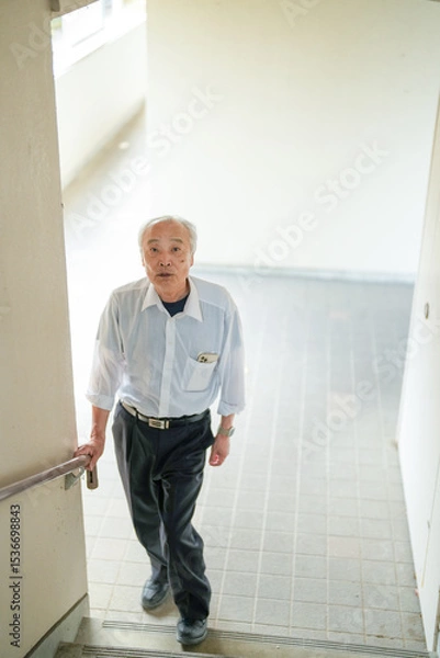 Fototapeta In Japan, a Japanese man in his late 70s, wearing a white shirt, climbs the stairs of an apartment using the handrail for support. This peaceful moment highlights aging with dignity and the quiet stre