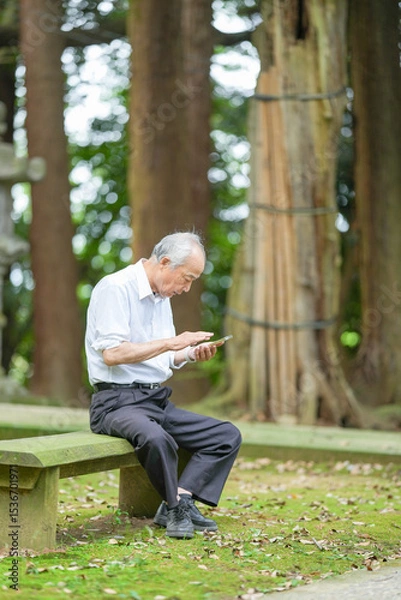 Fototapeta In rural Japan, a Japanese man in his late 70s sits at a peaceful countryside shrine, wearing a white shirt and hat. He uses his smartphone, surrounded by lush greenery in the early summer sunlight.