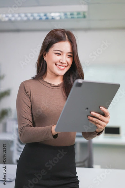 Fototapeta Young businesswoman is smiling while using a digital tablet in a modern office, showcasing the efficiency and connectivity of technology in the workplace