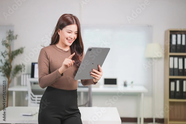 Fototapeta Happy Asian businesswoman standing in a modern office, using a digital tablet while pointing at the screen, smiling confidently and engaging in her work
