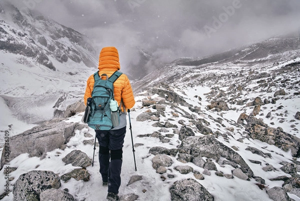Fototapeta Hiker with backpack walks along snowy trail through mountain pass against backdrop of an approaching snow storm in Himalayas.